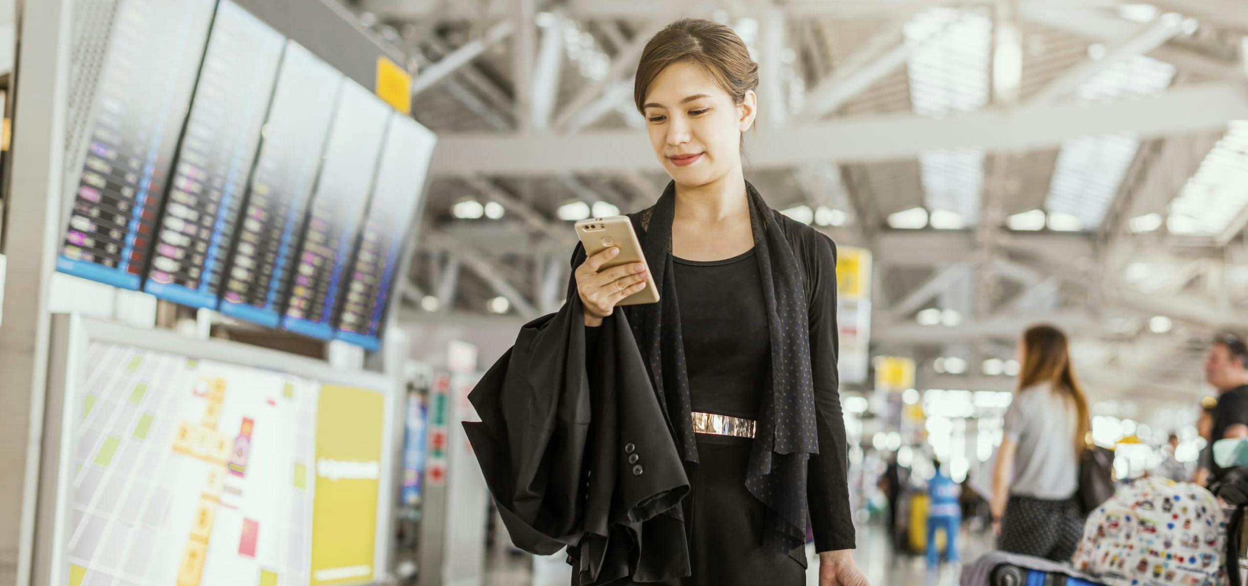 Woman in business clothes walking through an airport looking at her phone and holding a jacket.